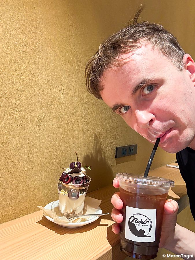 Marco Togni beve da un bicchiere con il logo Naki vegan sweets accanto a un dessert al cucchiaio nel locale di Namba.