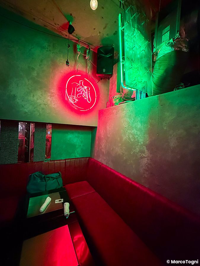 Interno a Neo Shinjuku Atsushi con luci neon verdi e rosse che illuminano un divanetto in pelle rossa.