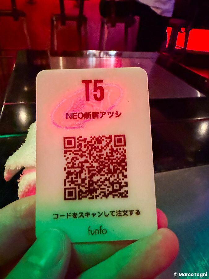 Una mano tiene una tessera bianca con codice QR per ordinare a Neo Shinjuku Atsushi, in un interno illuminato al neon.