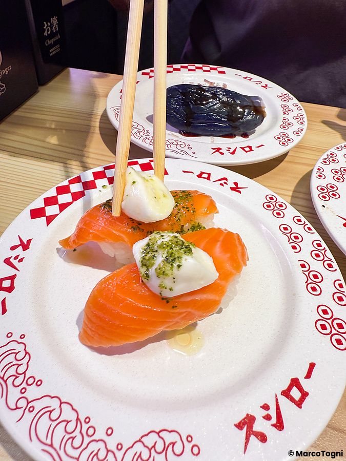 Bacchette sollevano un nigiri di salmone con salsa sopra un piatto brandizzato del ristorante Sushiro.