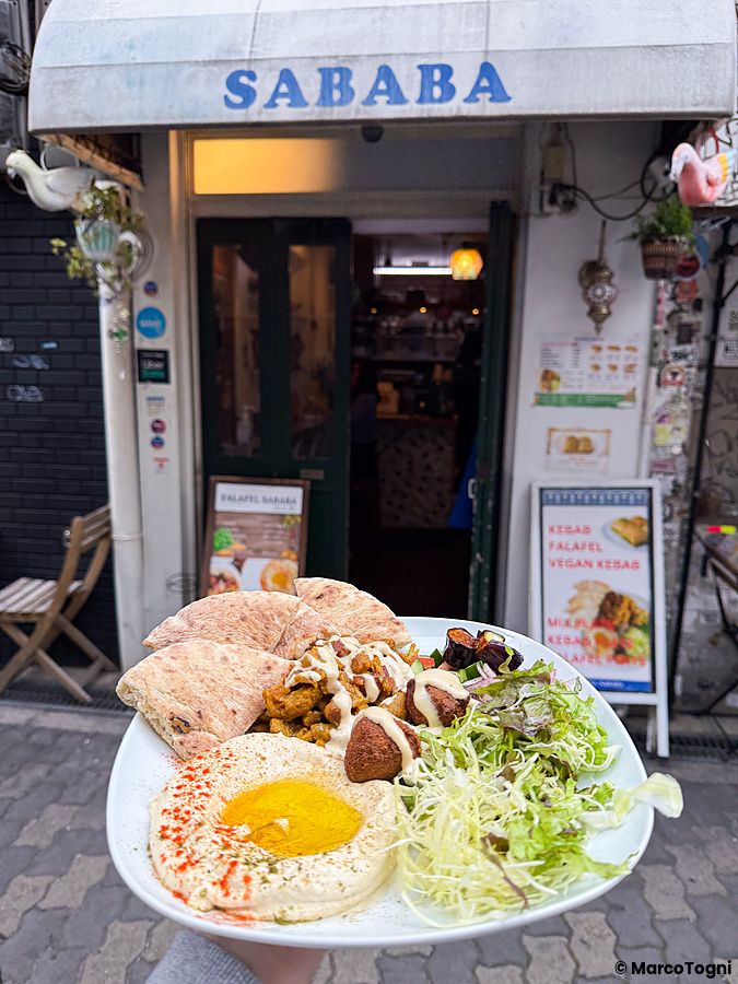 Un piatto misto vegano con falafel e pane pita in primo piano davanti all'ingresso del ristorante Falafel Sababa.