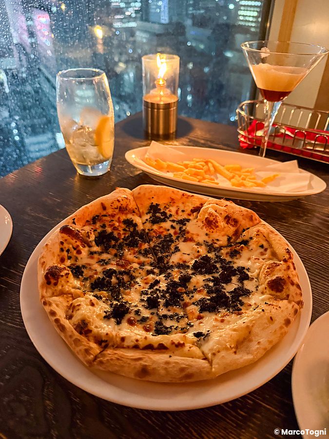 Una pizza su un piatto al Cafe Legato a Shibuya, con una finestra che mostra lo skyline notturno della città.