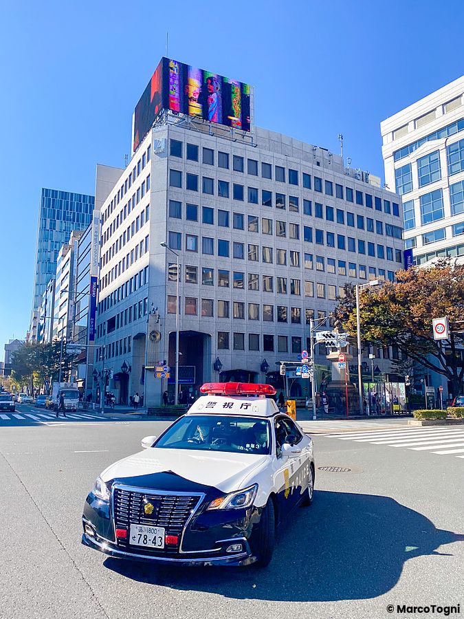 Auto della polizia ferma a un incrocio in una strada di Tokyo con edifici moderni sullo sfondo.