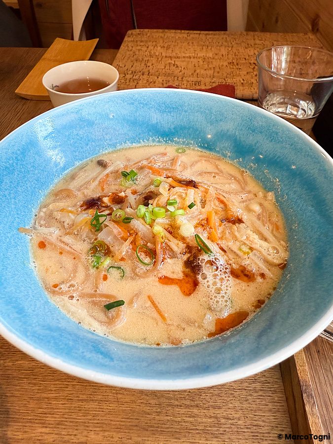 Una ciotola blu di ramen vegano con verdure e cipollotto servita su un tavolo di legno presso il Cafe Prana.