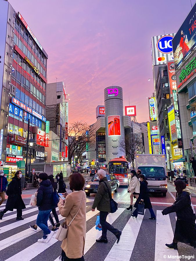 Persone attraversano le strisce pedonali di Shibuya al tramonto con il centro commerciale Shibuya 109 sullo sfondo.