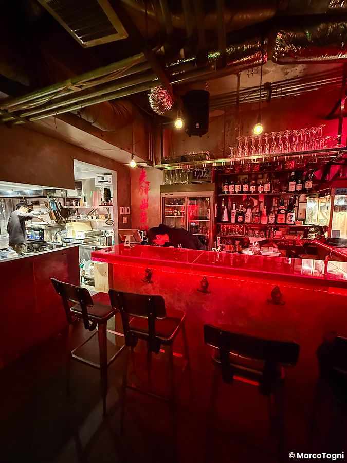 Interno a Neo Shinjuku Atsushi con bancone rosso, sgabelli in metallo e cucina a vista in stile cyberpunk.