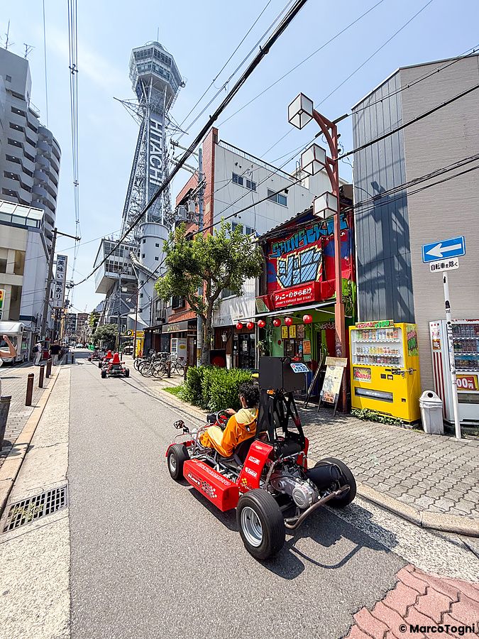 Un go-kart rosso guidato da una persona percorre una strada a Shinsekai, Osaka, con la Tsutenkaku Tower sullo sfondo.
