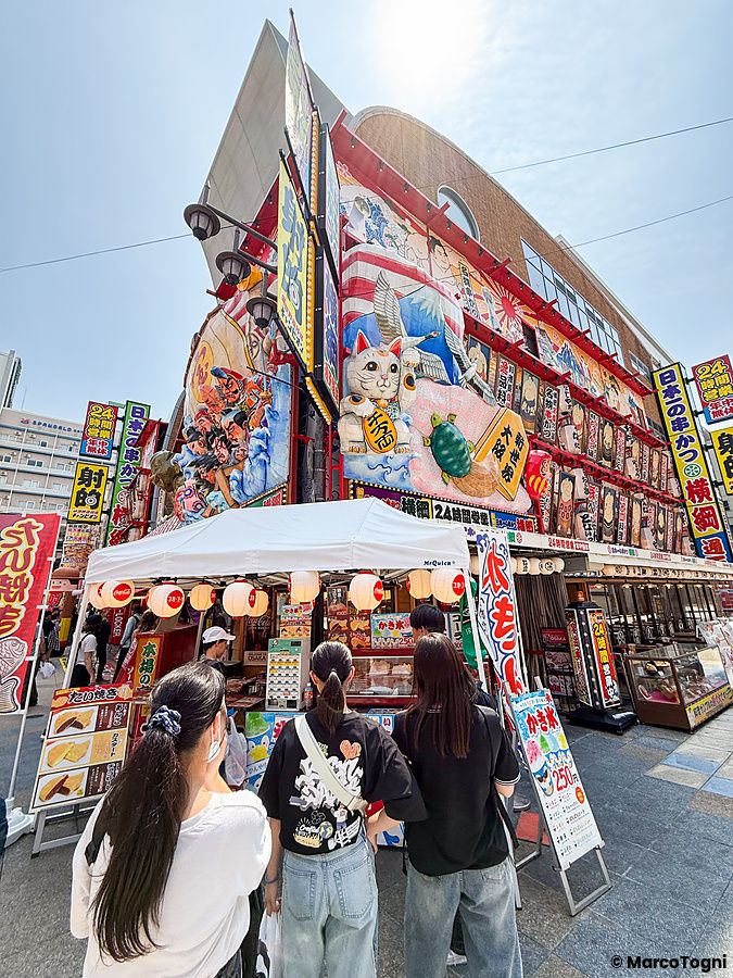 Tre persone osservano un banco di street food davanti a un edificio decorato nel quartiere di Shinsekai a Osaka.