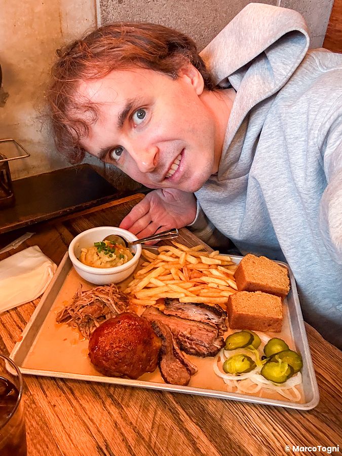 Marco Togni in felpa grigia scatta un selfie accanto a un vassoio di carne barbecue servito presso il ristorante Smokehouse.
