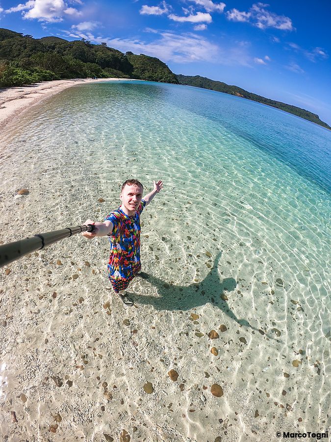 Marco Togni in piedi nelle acque trasparenti della spiaggia di Ida a Okinawa, ripreso in un selfie con un bastone.