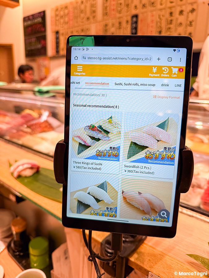 Un tablet per ordinare a Uogashi Nihonichi mostra il menù digitale con foto di sushi sopra il bancone del ristorante.