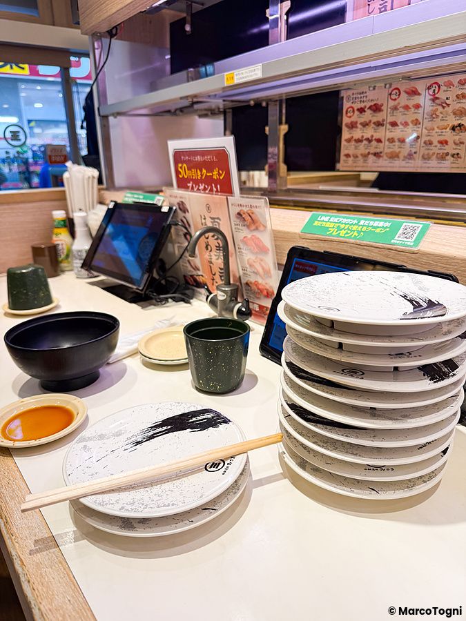 Pila di piatti usati su un tavolo di Kura Sushi a Tokyo, con bacchette e schermi per ordinare il cibo.