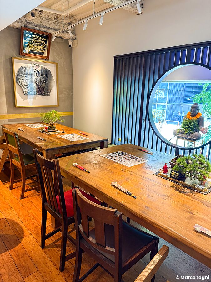 Interno del Tokyo Vegan Ramen Center ad Harajuku con tavoli in legno, bonsai, una giacca in cornice e una finestra tonda.