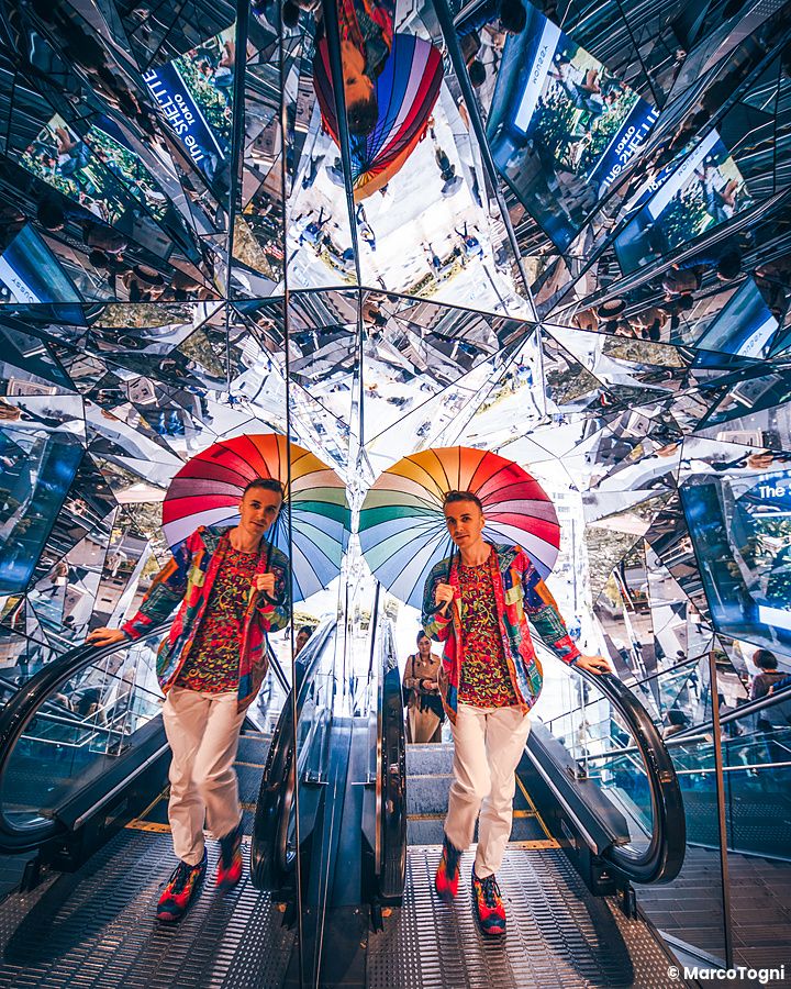 Marco Togni con un ombrello colorato su una scala mobile circondata da specchi al Tokyu Plaza ad Harajuku.