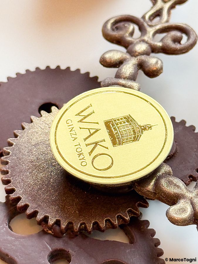 Dettaglio del logo su ingranaggi di cioccolato da mangiare a Wako Tea Salon con una placca dorata circolare.