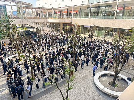 Una numerosa coda per prendere l'autobus composta da molte persone in una piazza pavimentata davanti a edifici moderni.