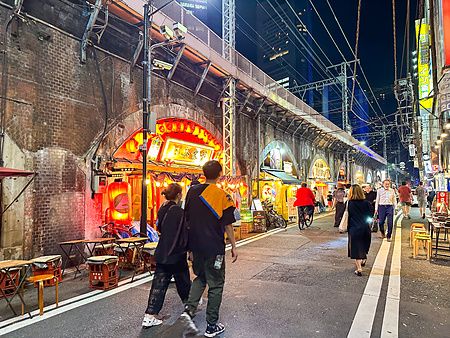 Persone camminano di notte lungo una strada sotto i viadotti in mattoni di Shimbashi a Tokyo, illuminata da insegne luminose.