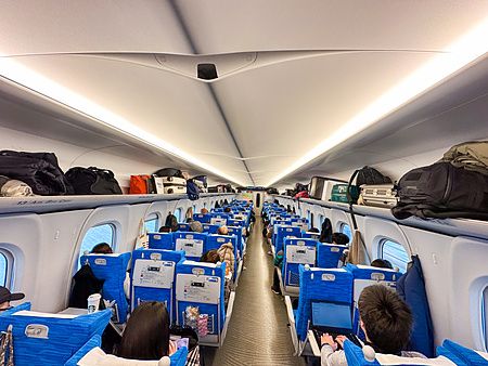 Interno di una carrozza passeggeri sul treno shinkansen con sedili blu occupati da persone e bagagli negli scomparti.