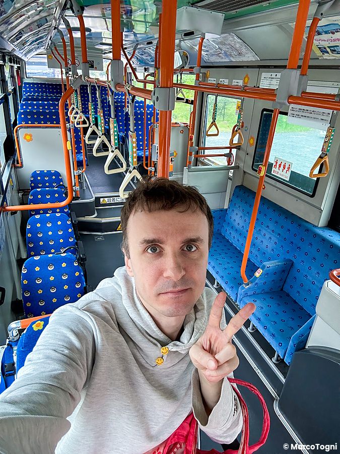 Marco Togni si scatta un selfie all'interno di un autobus di Tokyo con sedili blu e maniglie arancioni.