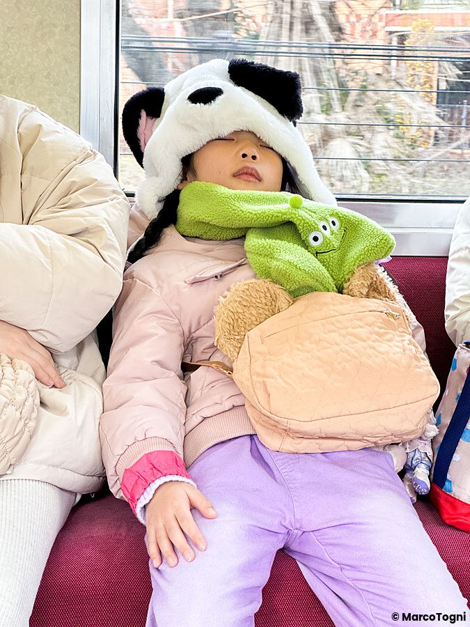 Una bambina dorme sul sedile di un treno in Giappone, indossando un cappello da panda e una sciarpa verde.
