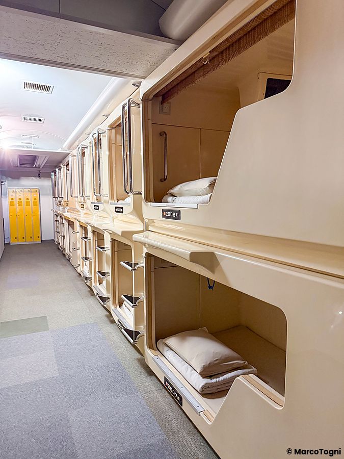 Lungo corridoio di un Capsule Hotel in Giappone con moduli letto sovrapposti color crema e armadietti gialli in fondo.