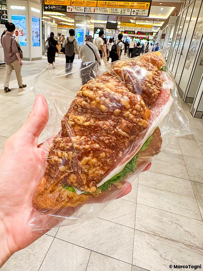 Un croissant salato con prosciutto crudo tenuto in mano davanti al passaggio pedonale di una stazione a Tokyo da Paul.
