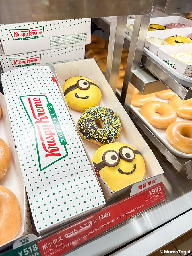 Una scatola con tre ciambelle a tema Minions e logo Krispy Kreme esposta in una vetrina insieme ad altre ciambelle.