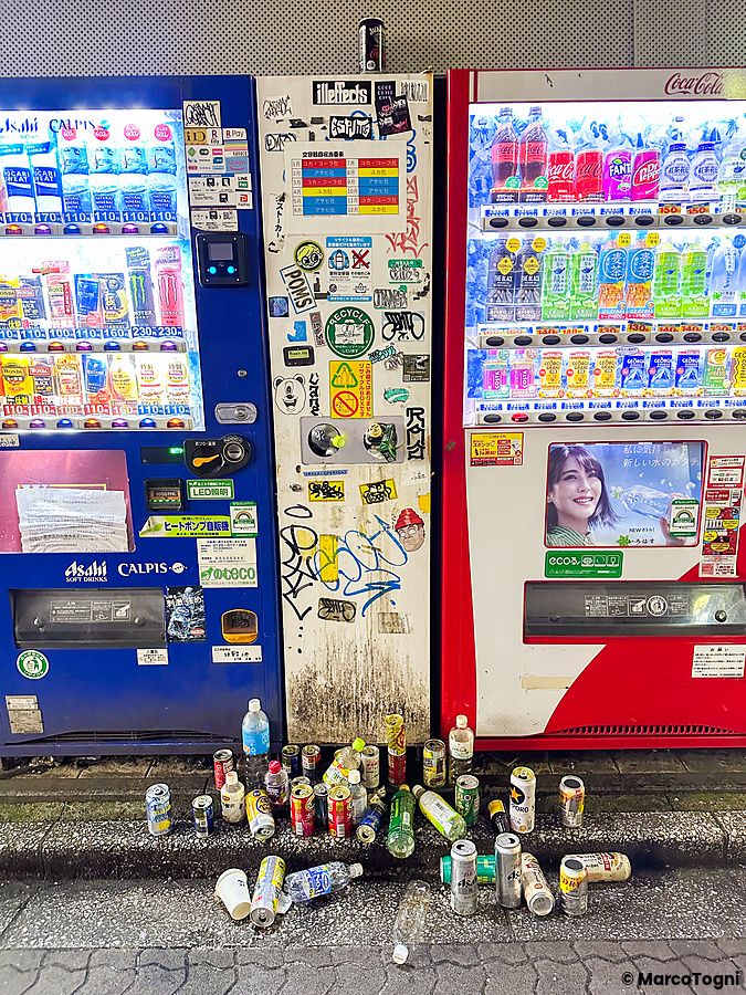 Distributori automatici di bevande a Tokyo circondati da rifiuti e lattine vuote a terra in una zona urbana degradata.