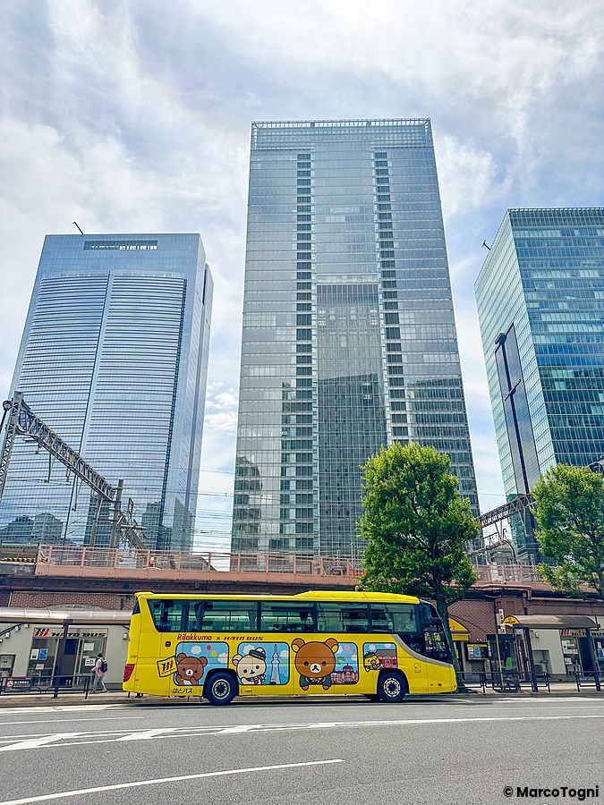 Un bus turistico Hato no Bus giallo con grafica di Rilakkuma sosta in strada davanti ai grattacieli vicino alla stazione d...
