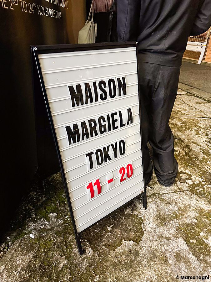 Cartello informativo della mostra di Maison Margiela a Tokyo con una persona in piedi accanto ad esso.