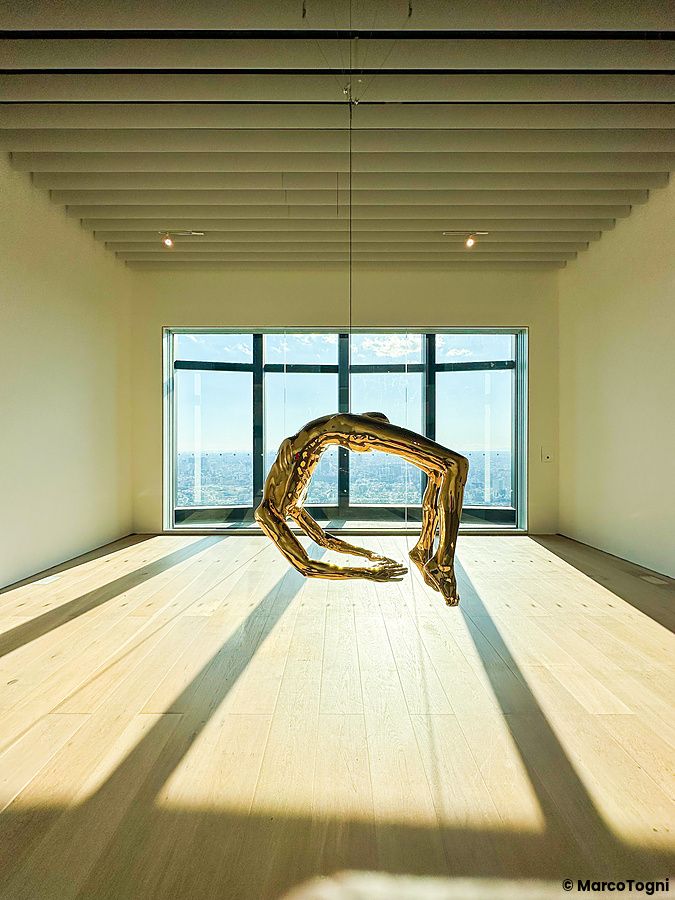 Una scultura dorata sospesa al centro di una sala del Mori Art Museum a Tokyo, con una vista panoramica della città.