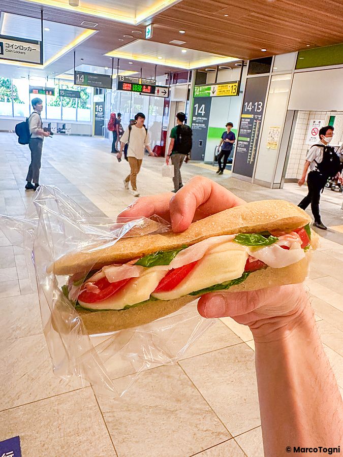 Un sandwich di pane PAUL tenuto in mano nella stazione di Shinjuku in Giappone, con passeggeri sullo sfondo.