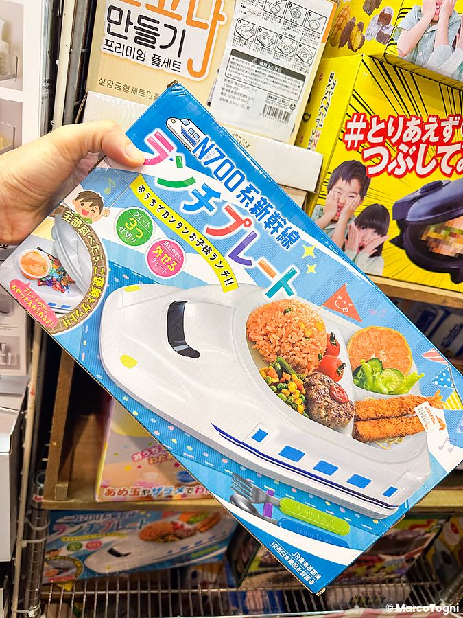 Una mano sostiene la scatola di un piatto per pranzo a forma di treno shinkansen in un negozio Village Vanguard.