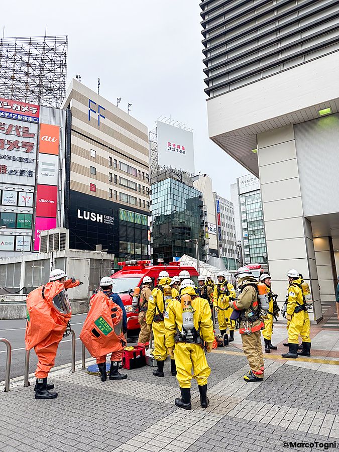 Gruppo di pompieri a Shinjuku con tute protettive gialle e arancioni riuniti su un marciapiede cittadino.