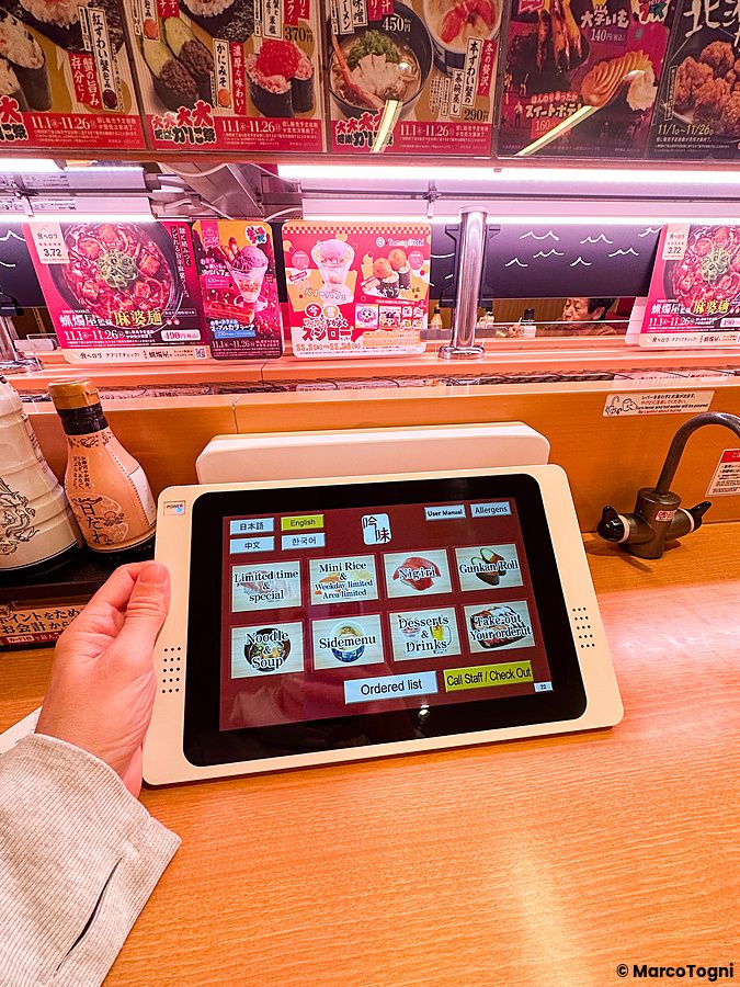 Una mano utilizza il tablet per ordinare a Sushiro, poggiato su un tavolo di legno in un locale luminoso.