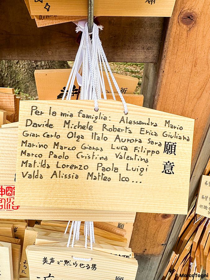 Una tavoletta di legno ema con scritte a mano in italiano appesa a un supporto in un tempio giapponese.
