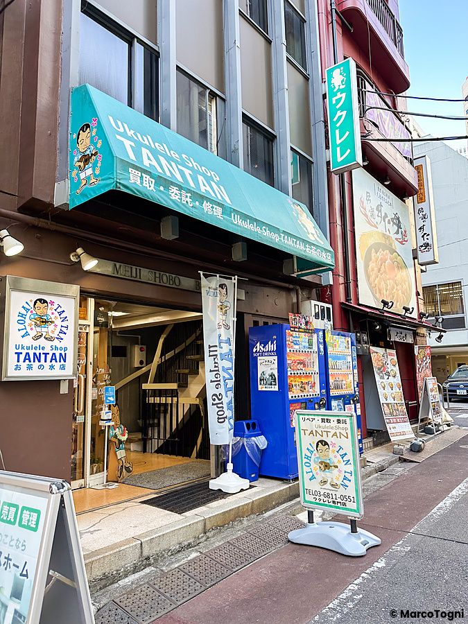 Esterno dello Ukulele shop Tantan ad Ochanomizu con tenda verde, cartelli promozionali e distributori automatici.