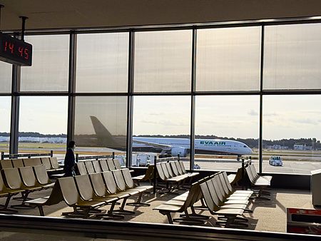 Veduta interna di una sala d'attesa all'aeroporto di Narita con un aereo Eva Air visibile attraverso una grande vetrata.