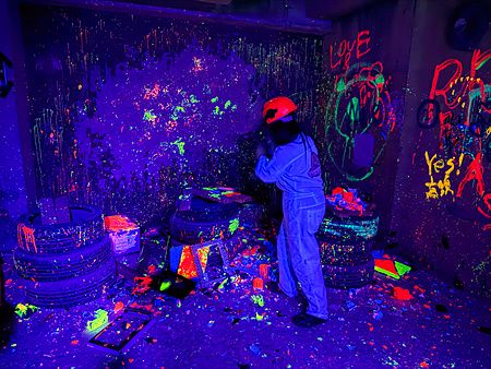 Una persona con tuta e casco distrugge oggetti nella rage room a Reeast Room, in un ambiente illuminato da luci UV.
