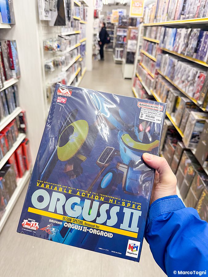 Una mano regge una scatola di un action figure Orguss II tra gli scaffali del negozio amiami Akihabara Figure Tower.