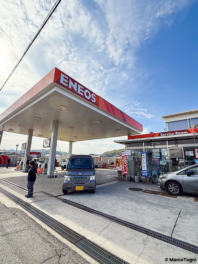 Un benzinaio Eneos in Giappone con una persona accanto a un'auto grigia sotto la pensilina rossa.