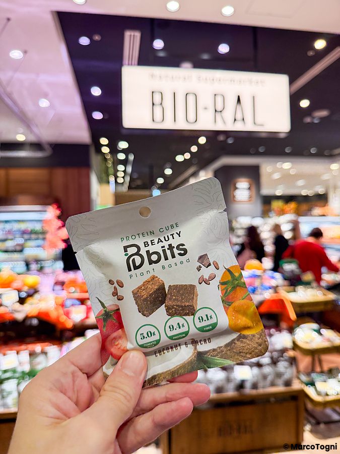 Una mano tiene una confezione di snack a Bio-ral con il logo del supermercato visibile sullo sfondo sfocato.