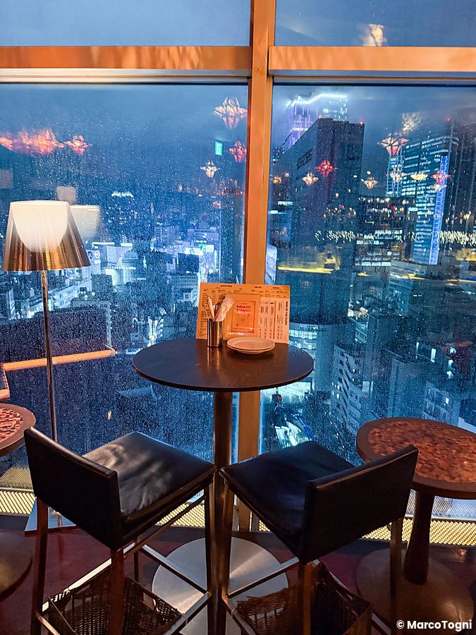Un tavolo rotondo con due sedie al Cafe Legato di Shibuya, con vista sulla città illuminata attraverso una vetrata.