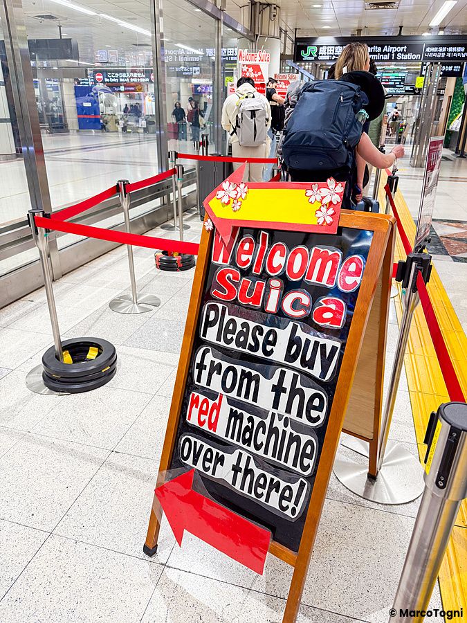 Cartello informativo per la carta Welcome Suica su cavalletto all'aeroporto di Narita con persone in fila sullo sfondo.