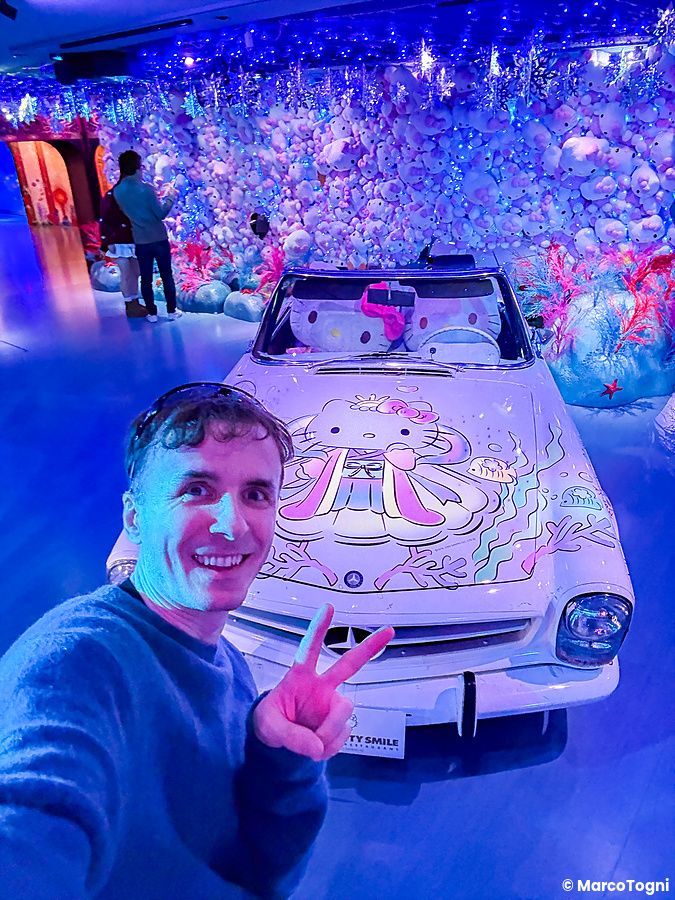 Marco Togni sorridente scatta un selfie davanti a un'auto personalizzata a tema Hello Kitty presso Hello Kitty Smile.
