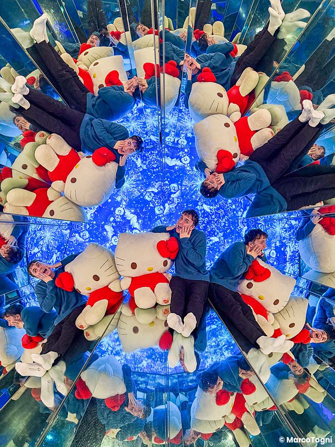 Marco Togni siede in una stanza a specchi presso Hello Kitty Smile sull'isola di Awaji, circondato da riflessi e peluche.