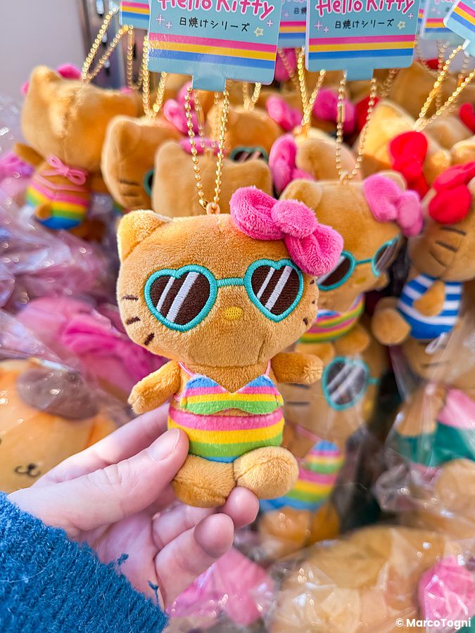 Una mano tiene un peluche di Hello Kitty abbronzata con occhiali a cuore e costume arcobaleno in un negozio.