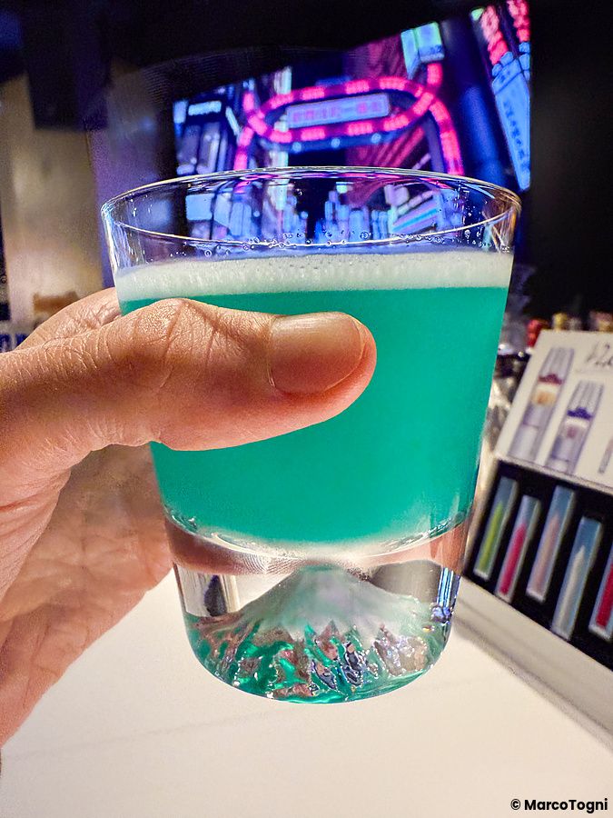 Una mano tiene un bicchiere con il rilievo del Monte Fuji alla base, riempito di un drink blu al Fujiyama Bar di Roppongi.
