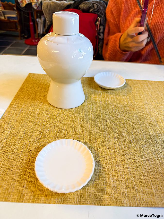 Un sakè di benvenuto al Yoshijin Battari Cafe con una caraffa in ceramica bianca e piattini su una tovaglietta senape.