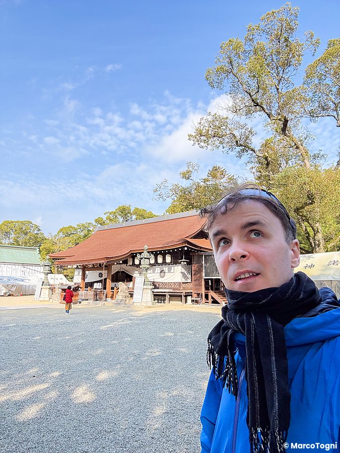 Marco Togni in primo piano al Santuario Izanagi con lo sfondo dell'edificio tradizionale in legno e alberi nel cortile.